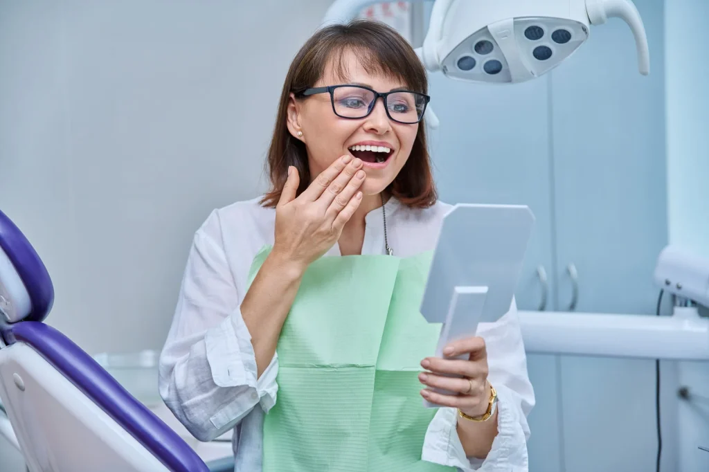 Smiling woman in dental office examining her teeth in mirror, showcasing satisfaction with dental treatment, highlighting dental implant success.