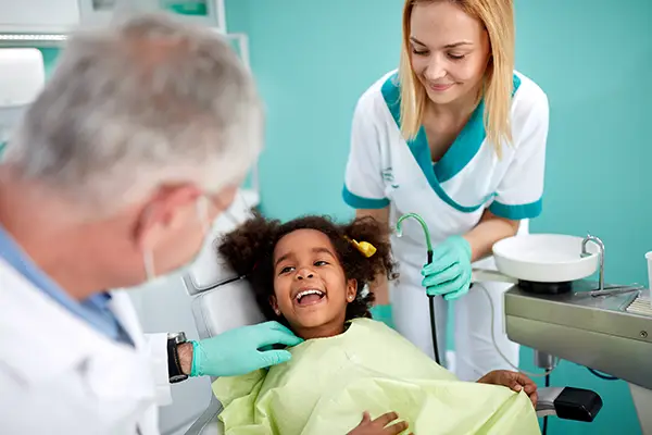 Child smiling in dental chair during routine checkup with dentist and hygienist at Palani Center for Dental Implants, emphasizing preventive oral care and comfort.