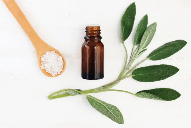 Brown glass essential oil bottle with sage leaves and a wooden spoon filled with salt, representing natural remedies in holistic dentistry.