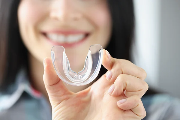 Woman smiling while holding a clear dental night guard, illustrating custom oral appliance for teeth grinding and TMJ relief.